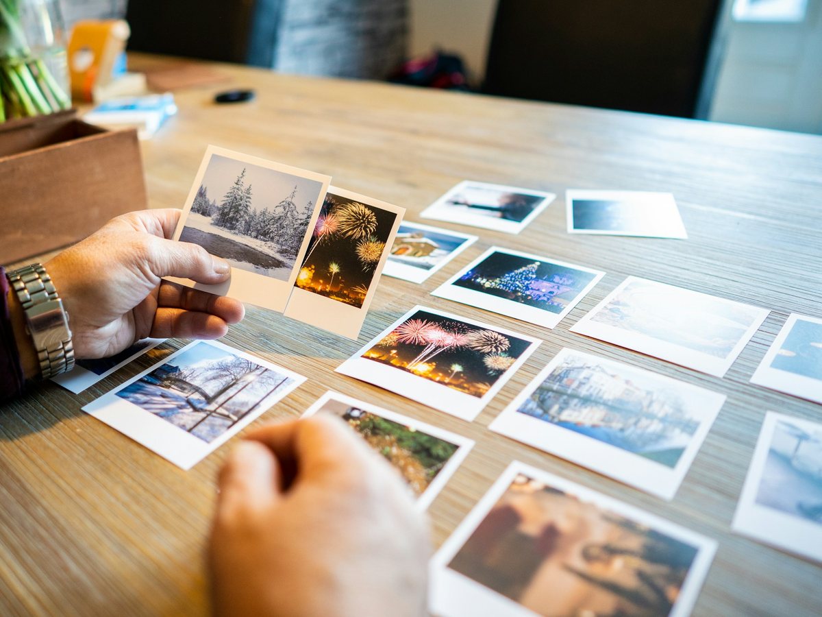 A hand holds two prints over a table of photographs laid out to choose from