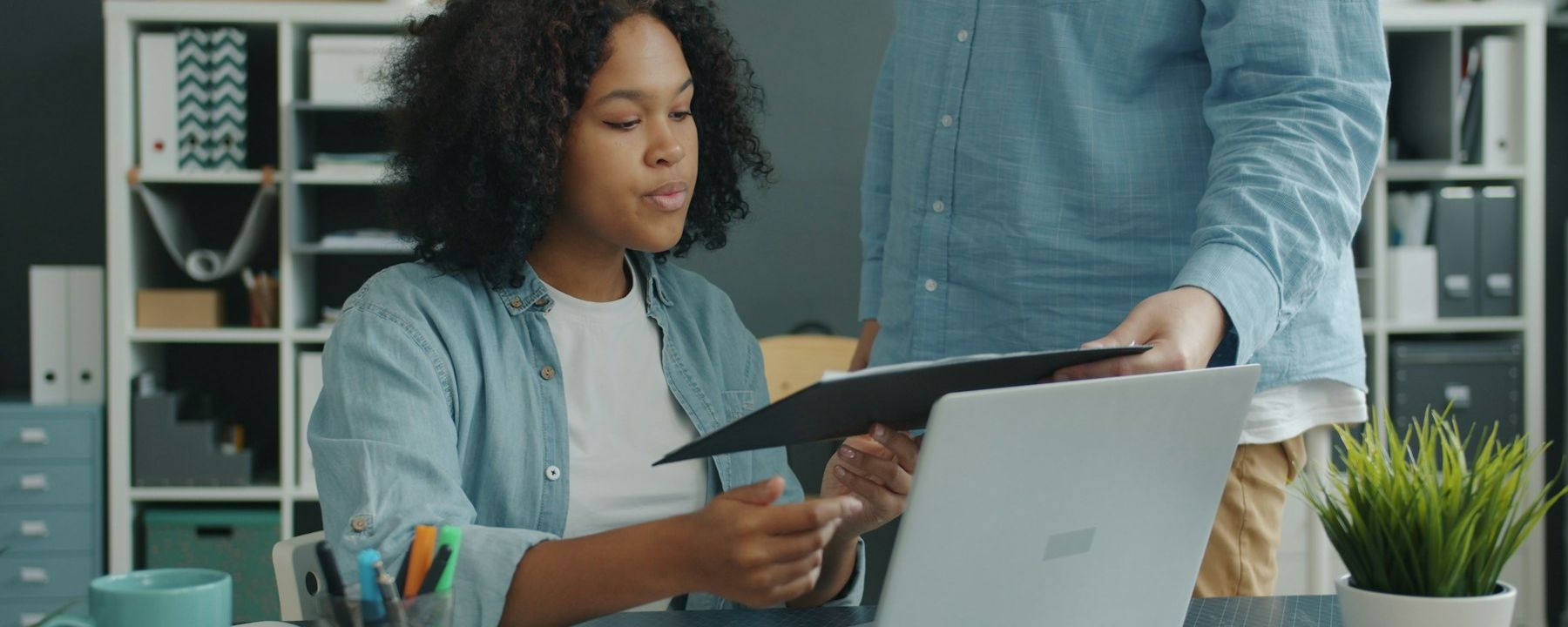 A woman at a laptop taking a printed document from a colleague standing beside her desk.