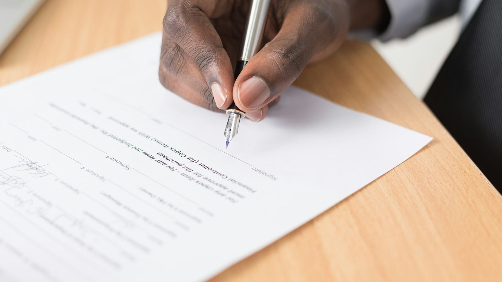 Close-up of a hand signing a printed form with a fountain pen on a light wooden desk.