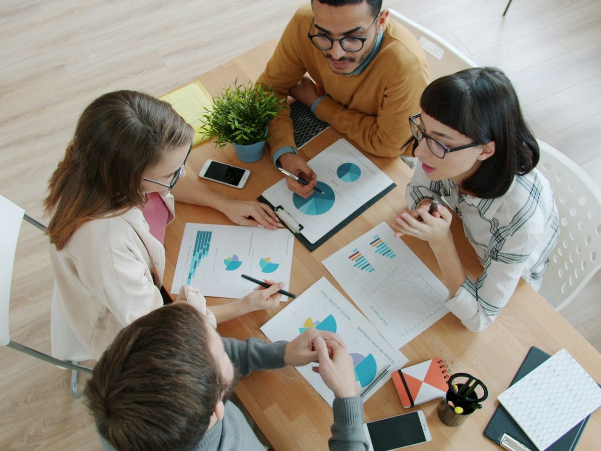Seen from above, four colleagues around a table covered in printed charts, one pointing at a pie chart.