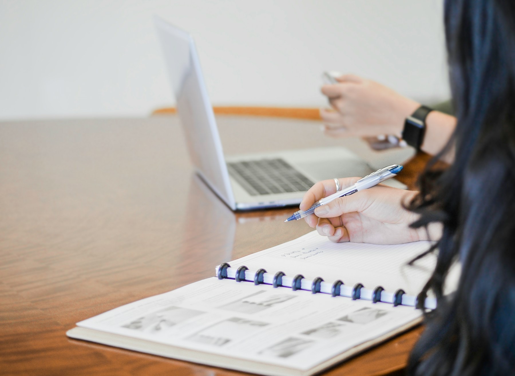A hand with a pen over an open notebook, a laptop on the meeting table behind it.