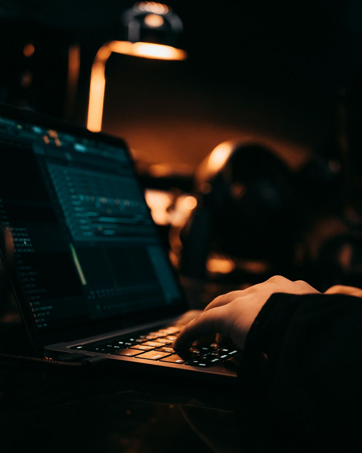 A person typing on a laptop in a dark room, code on the screen, a lamp behind.