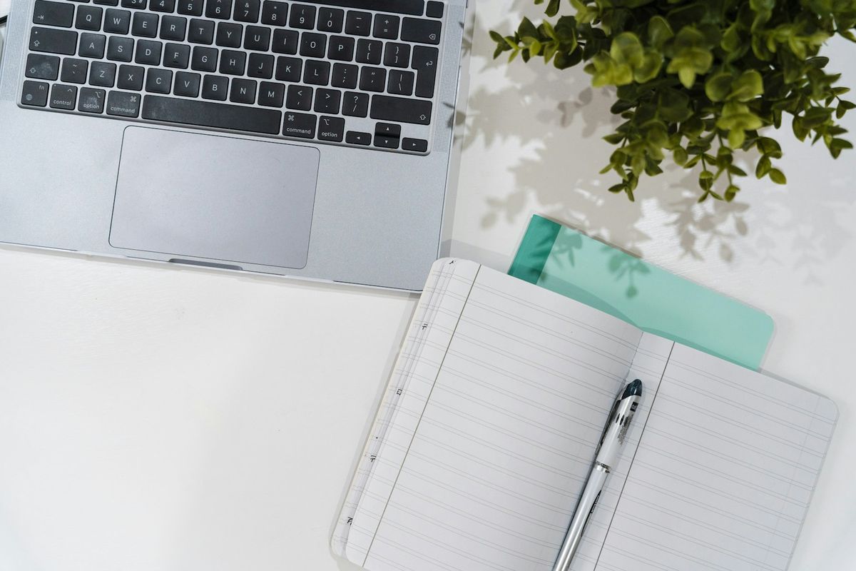 Seen from above, a laptop keyboard beside an open lined notebook with a pen lying on it, a small green plant at the corner of a white desk.