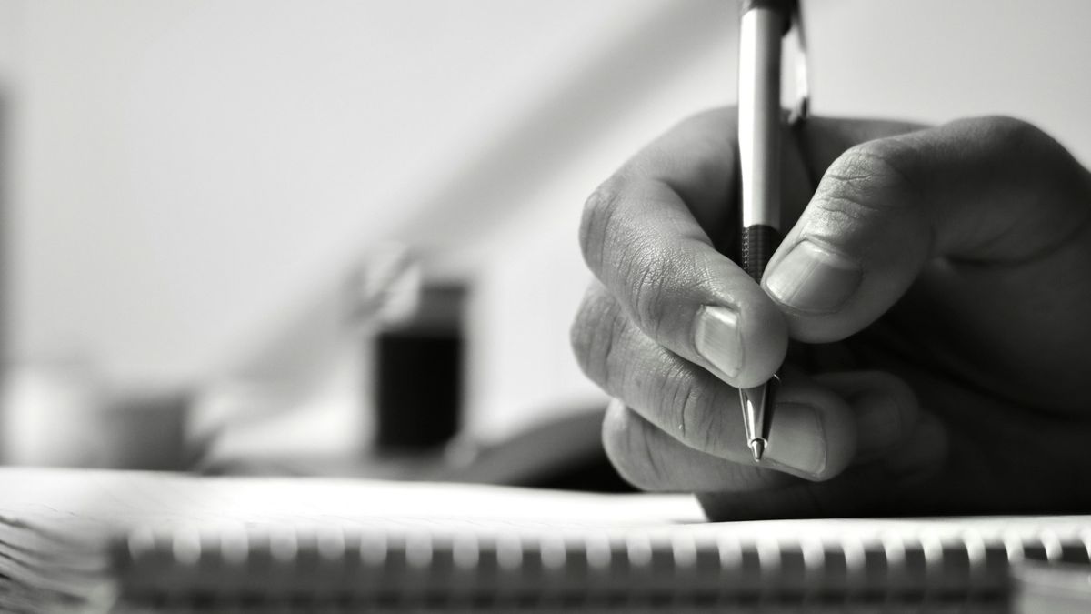 Black-and-white close-up of a hand holding a pen over a spiral-bound notebook.