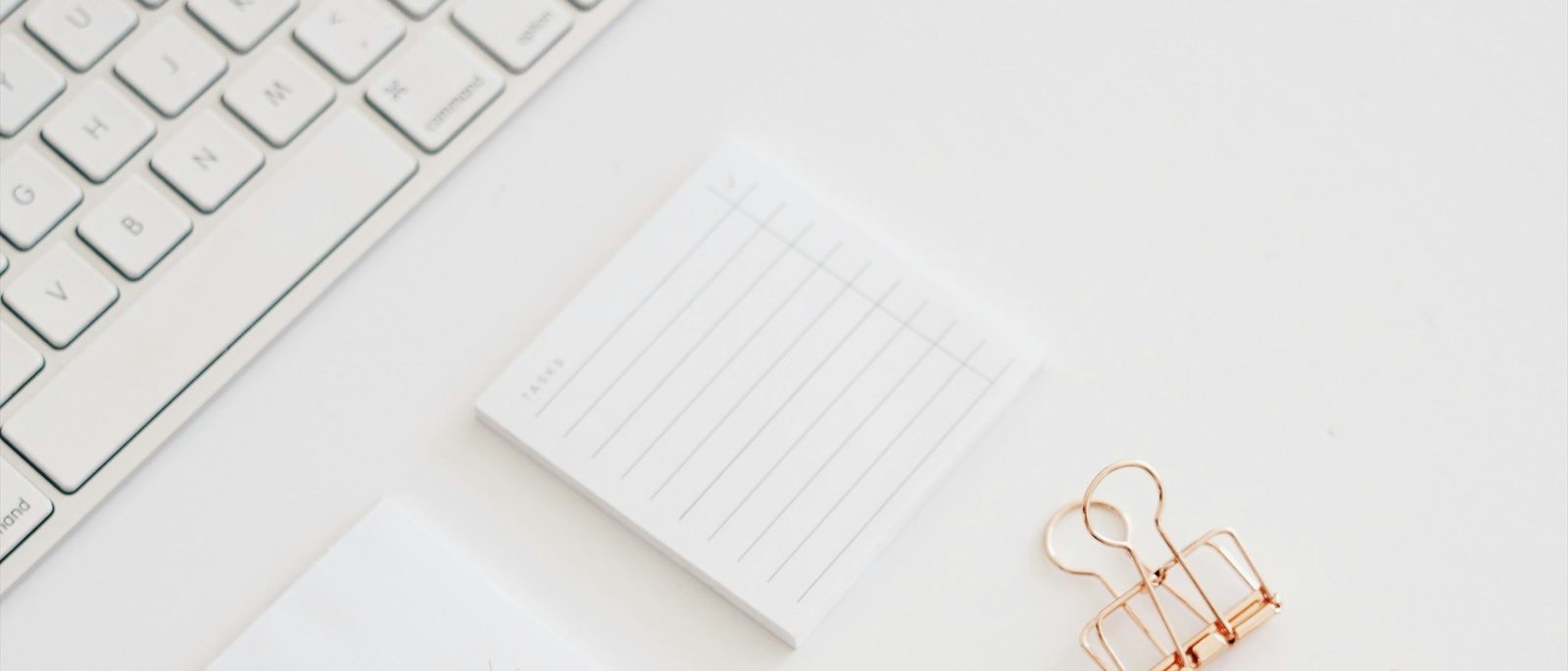 A small lined notepad and a binder clip on a white desk beside a keyboard