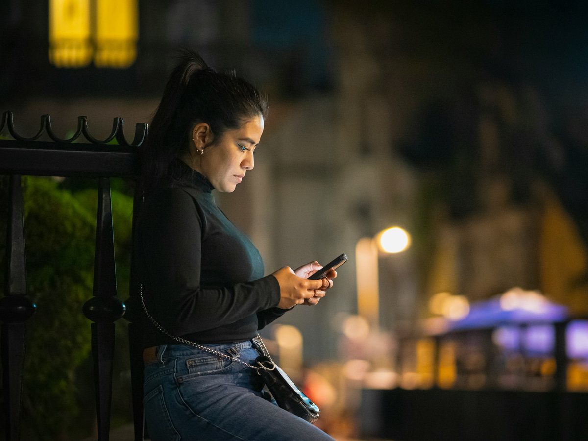 A woman sits by a railing on an evening street, reading her phone under warm city lights