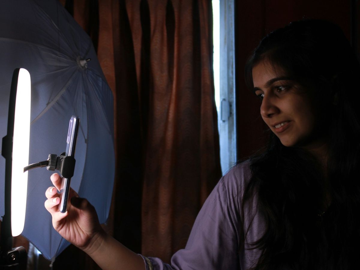A creator smiles at her phone on a stand beside a ring light in a dim room