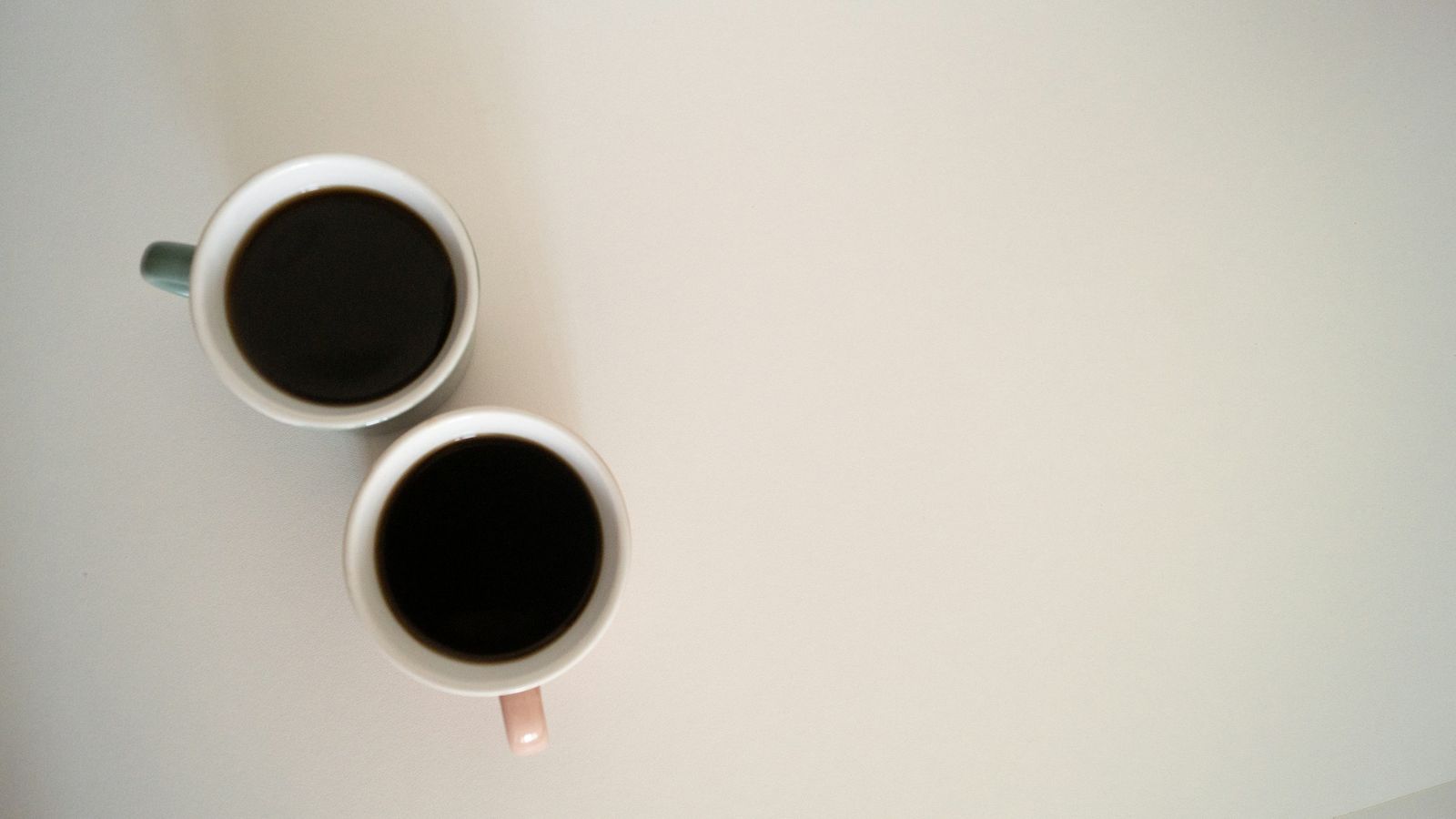 Two cups of black coffee side by side on a pale table, seen from above.