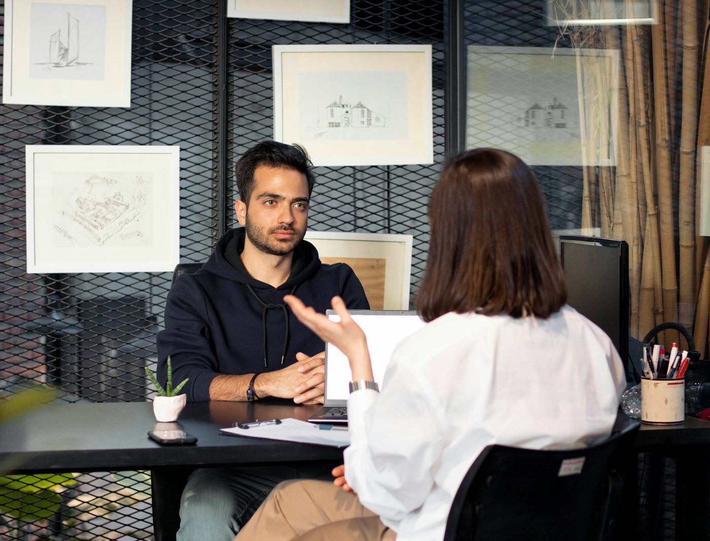 A man at a desk in conversation with a colleague across from him, framed drawings on the wall behind.