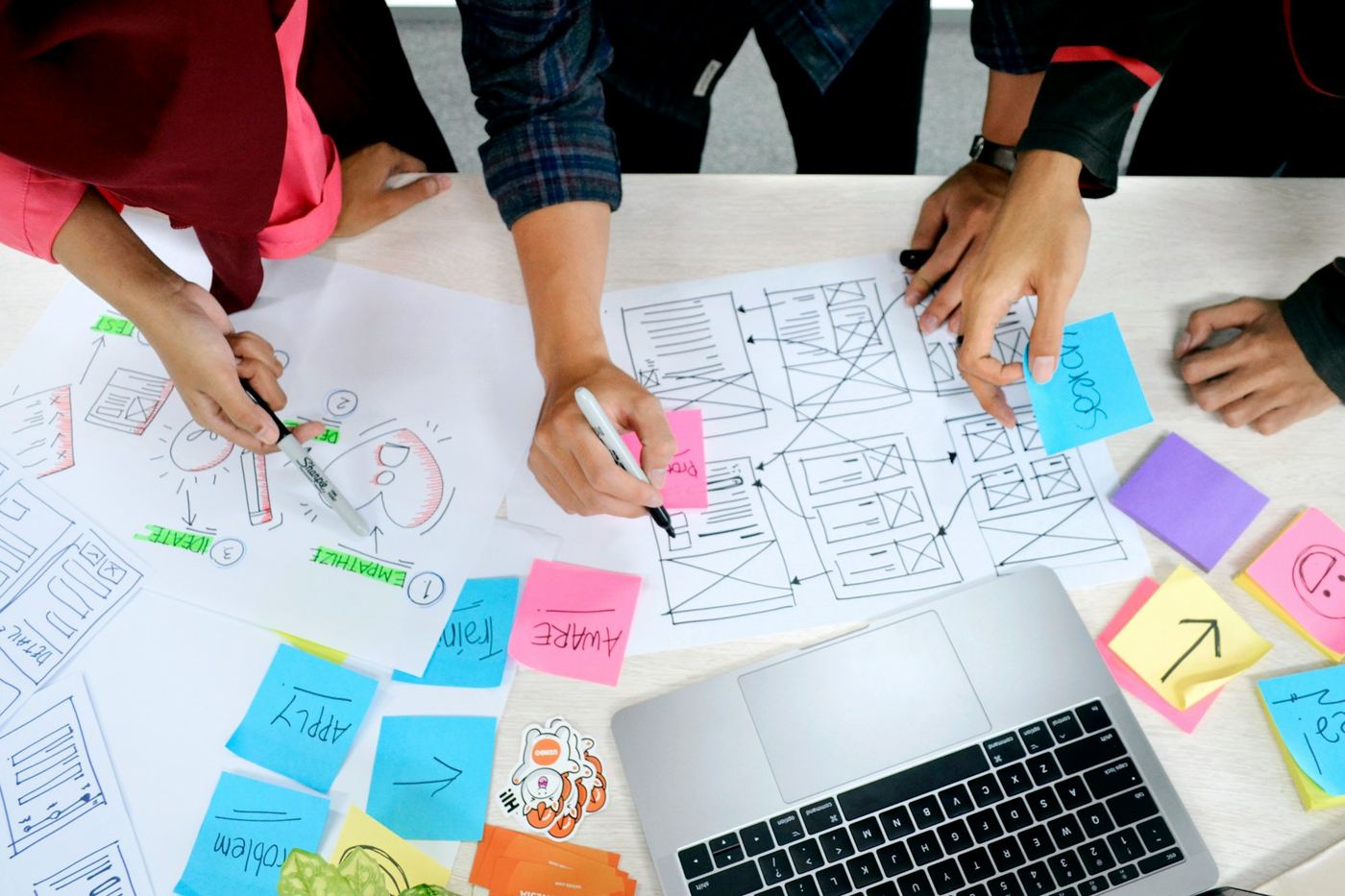 Hands arranging paper wireframes and coloured sticky notes on a table beside a laptop.