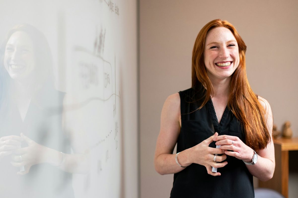 A woman smiling as she leads a session beside a whiteboard covered in a hand-drawn diagram.