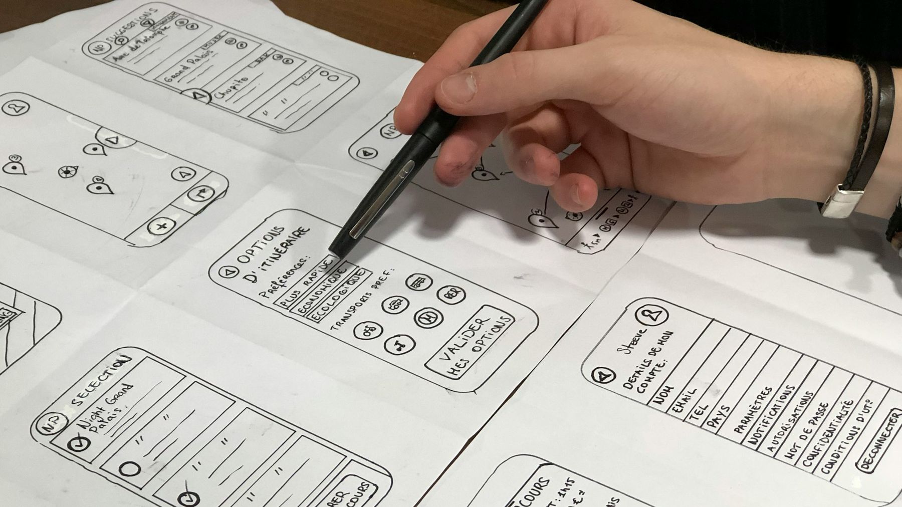 A hand pointing a pen at hand-drawn phone screens spread across a table: a map, a list of suggestions and a screen of route options and preferences.