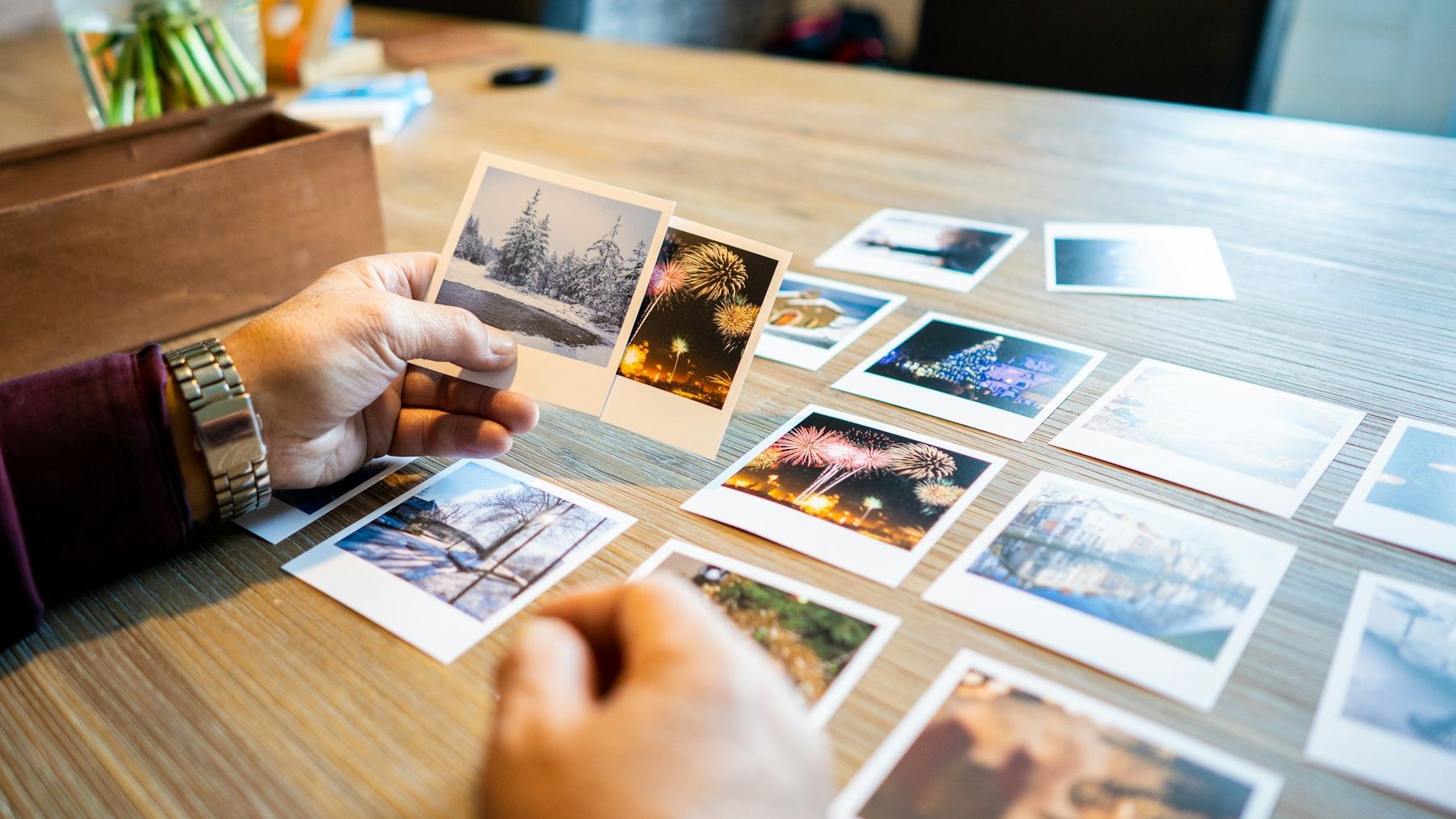 Hands sorting a spread of photographic prints on a wooden table, one print held up for a closer look.