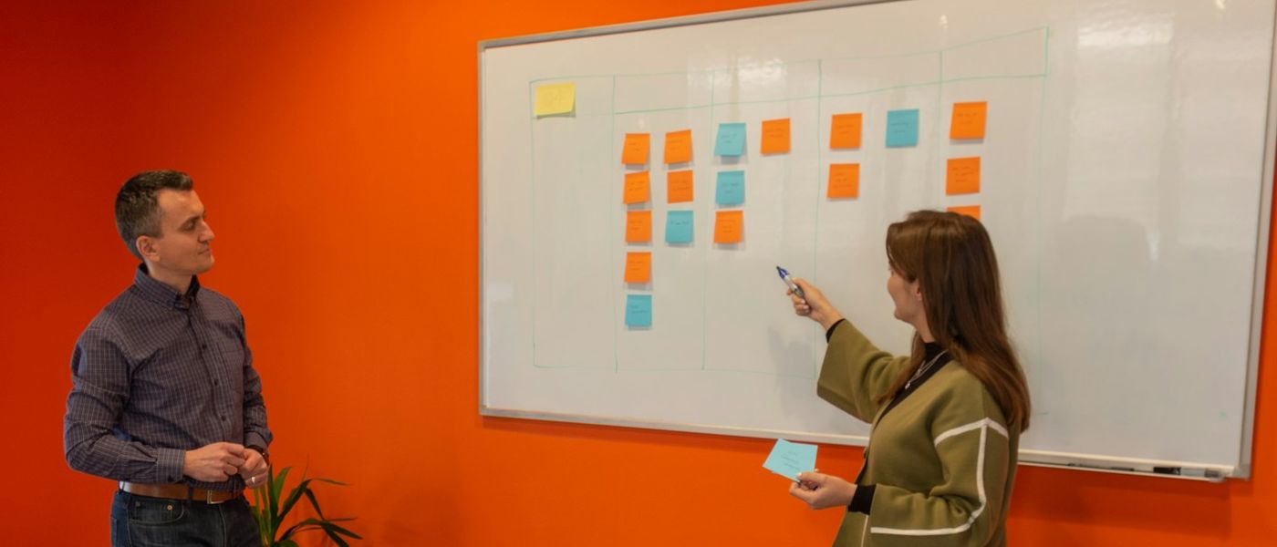 A woman pointing at a kanban board of sticky notes on a whiteboard while a colleague looks on, in a room with an orange wall.