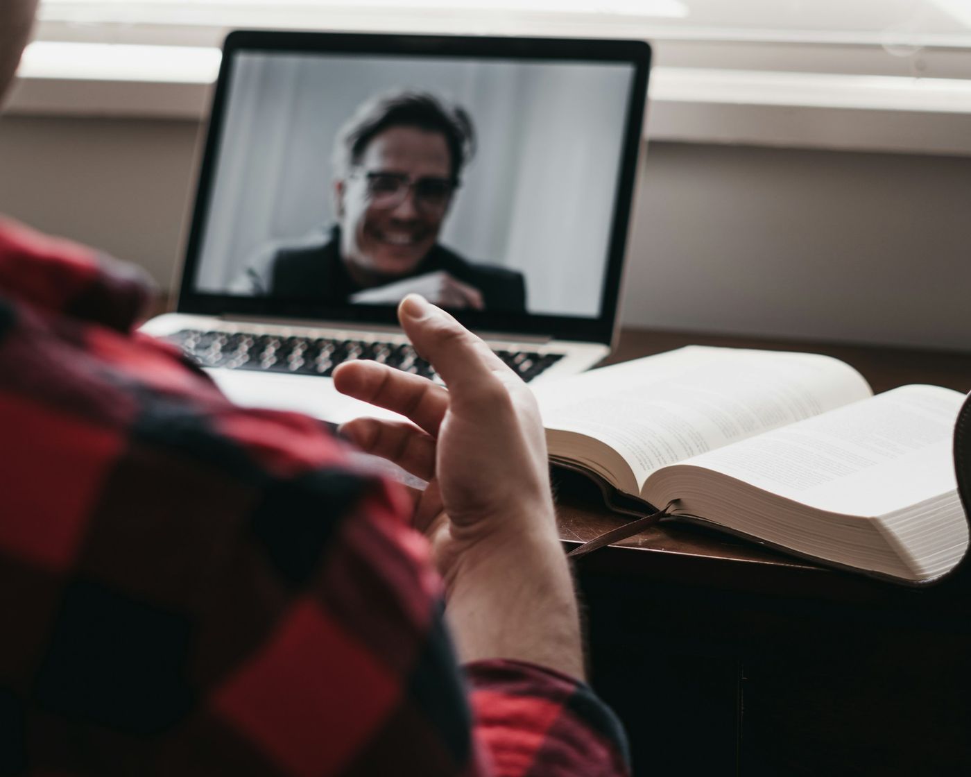 Over the shoulder of a person on a video call: a smiling man on the laptop screen, an open book on the desk.