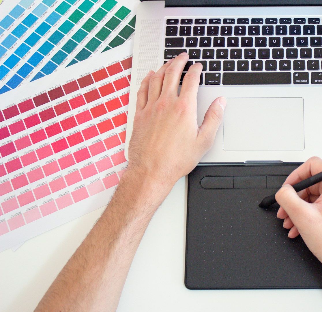 Hands at a laptop and a drawing tablet, beside printed sheets of colour swatches.
