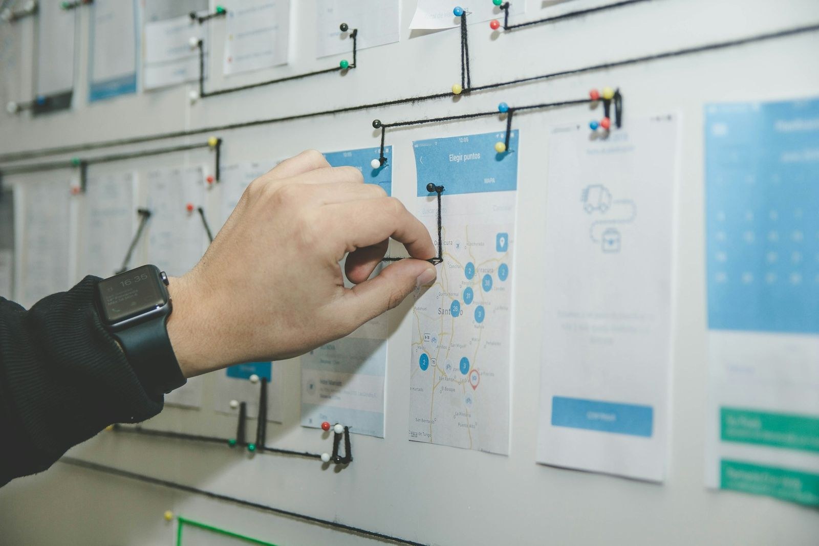 A hand pins string between printed app screens on a wall, mapping how one screen leads to the next.