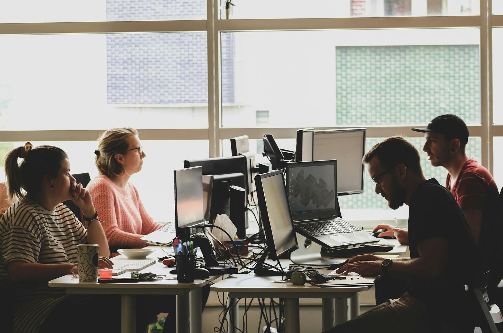 Four people at desks of monitors and laptops in a bright room with tall windows.