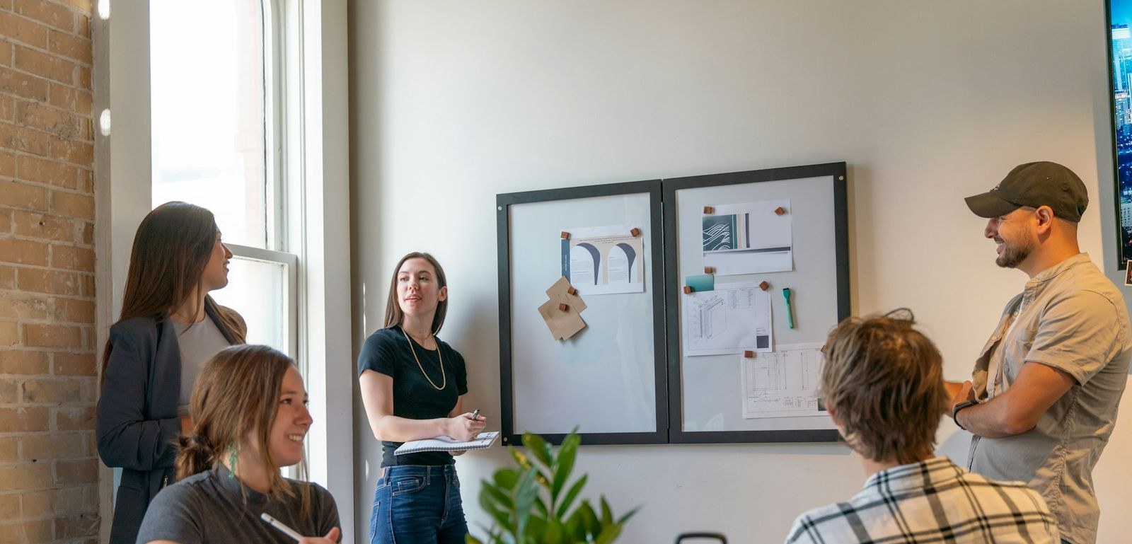 A team gathered at a pinboard in a sunlit room, one person presenting while the others take notes.