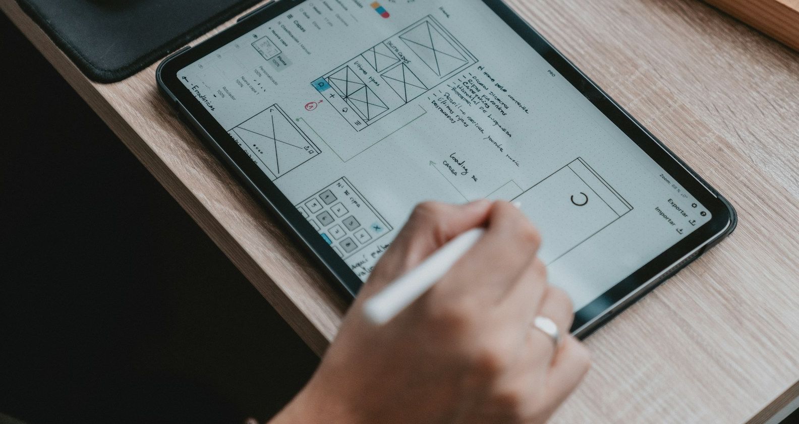 A hand with a stylus sketches screen layouts on a tablet resting on a wooden desk.