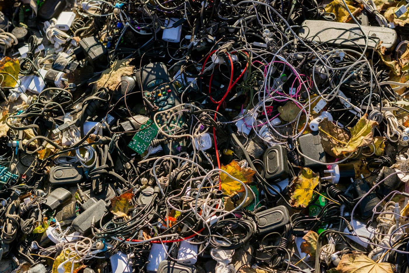 A heap of discarded chargers, cables, remote controls and circuit boards among dry leaves