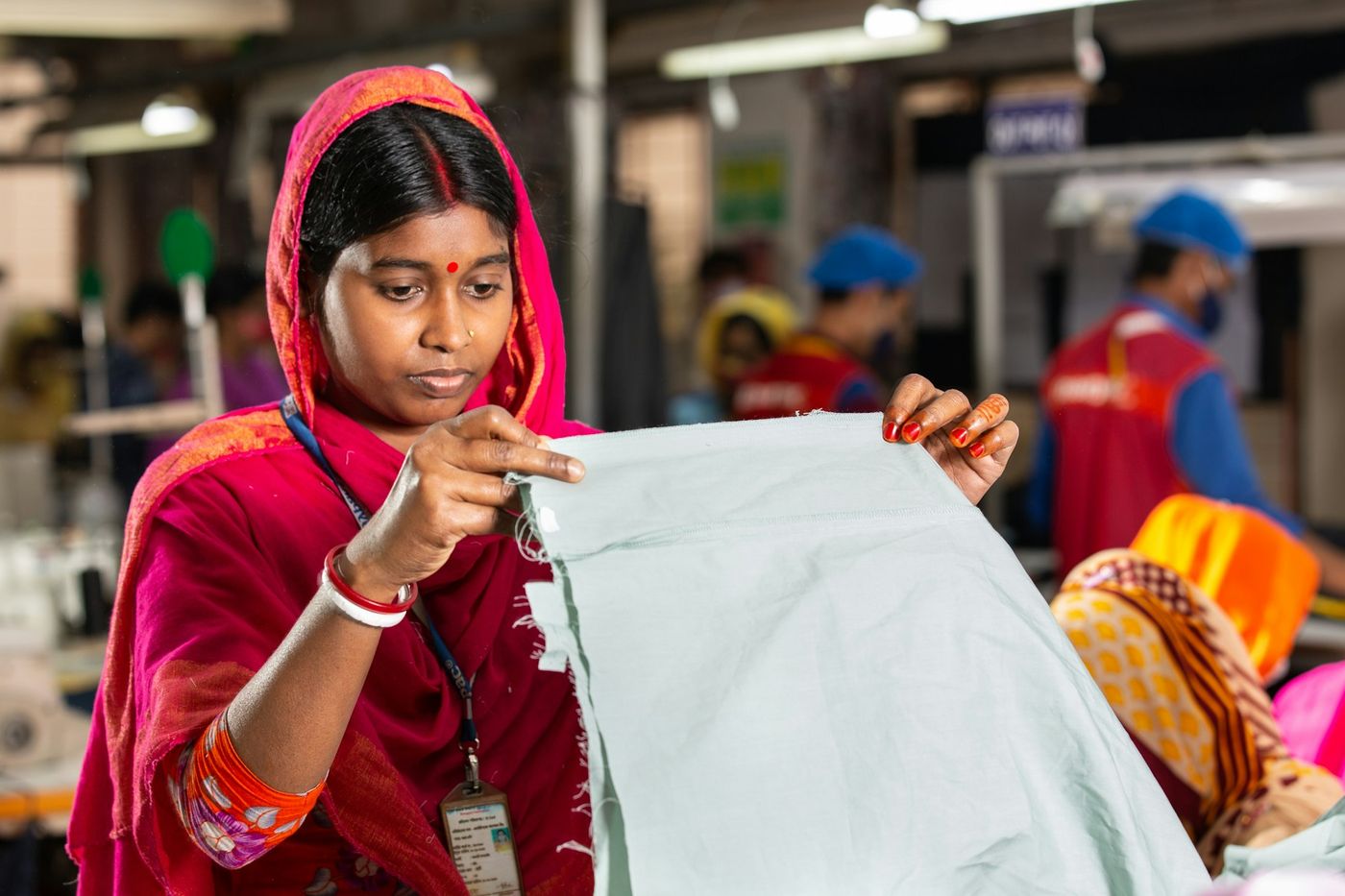 A garment worker holds up a pale green cut panel to inspect it, with sewing lines and colleagues behind her