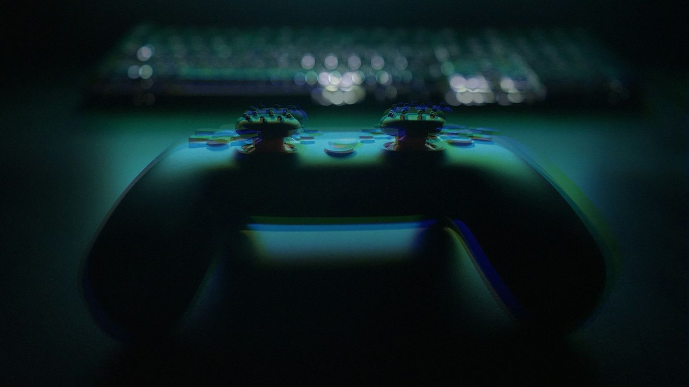 A game controller in deep shadow lit by teal light, with a keyboard glowing behind it