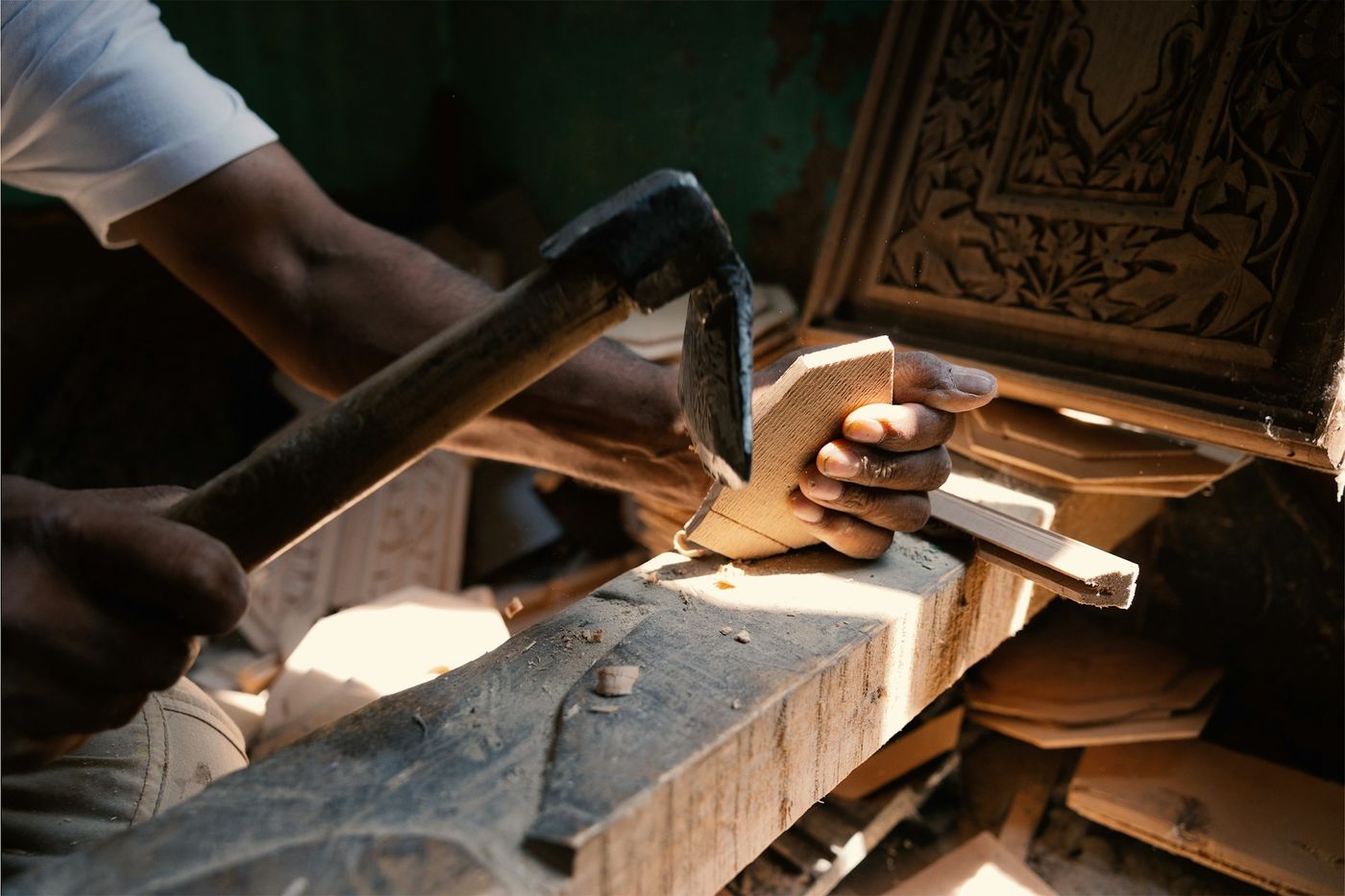A craftsman shapes a piece of wood with a hand adze beside a carved wooden panel