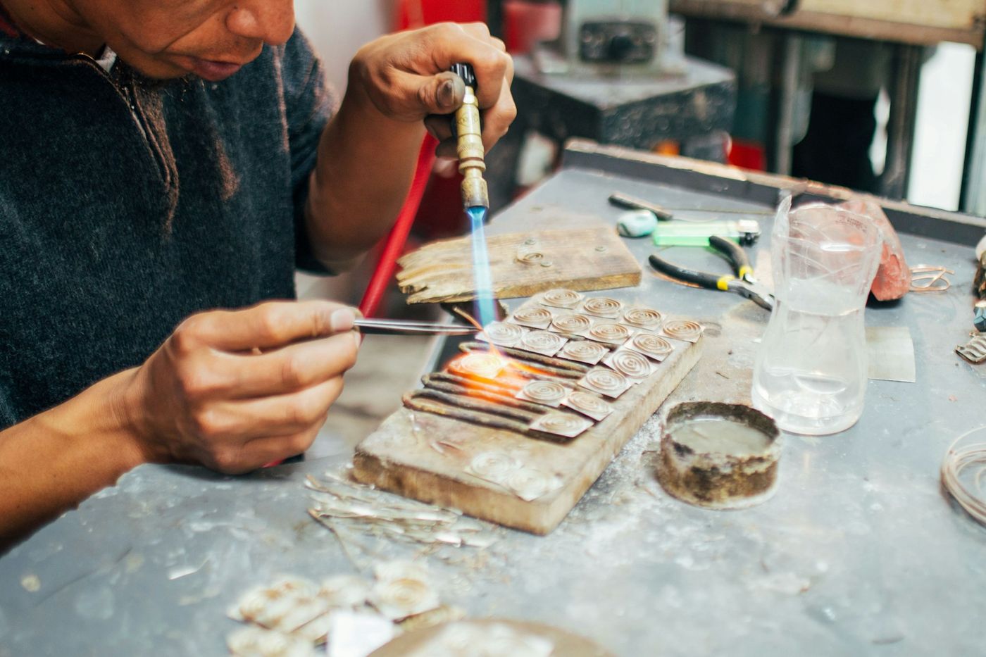 A craftsman heats small metal pieces with a fine torch on a cluttered jeweller’s bench