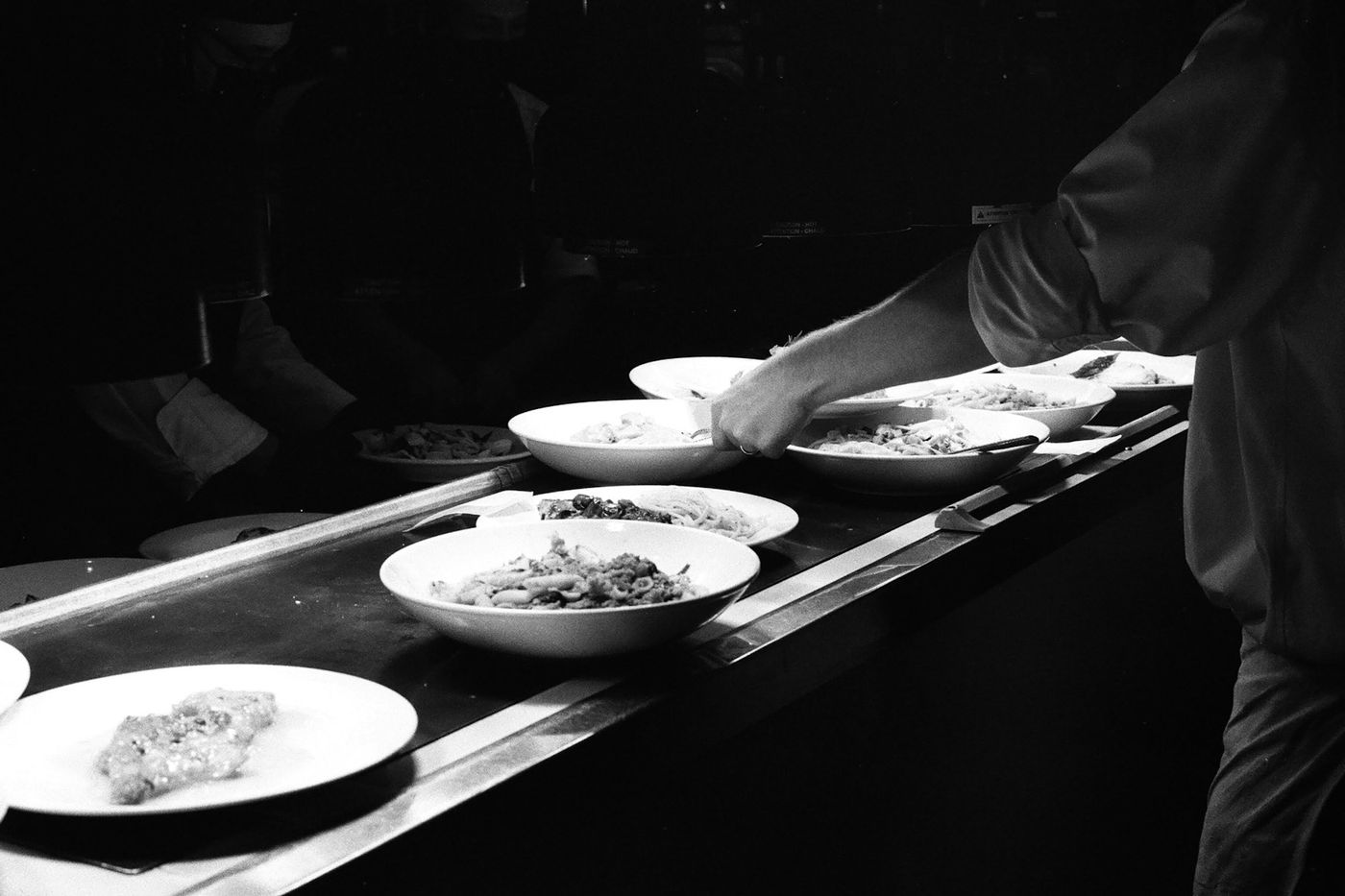 A black-and-white view of plated dishes lined up on a kitchen pass as a chef reaches across