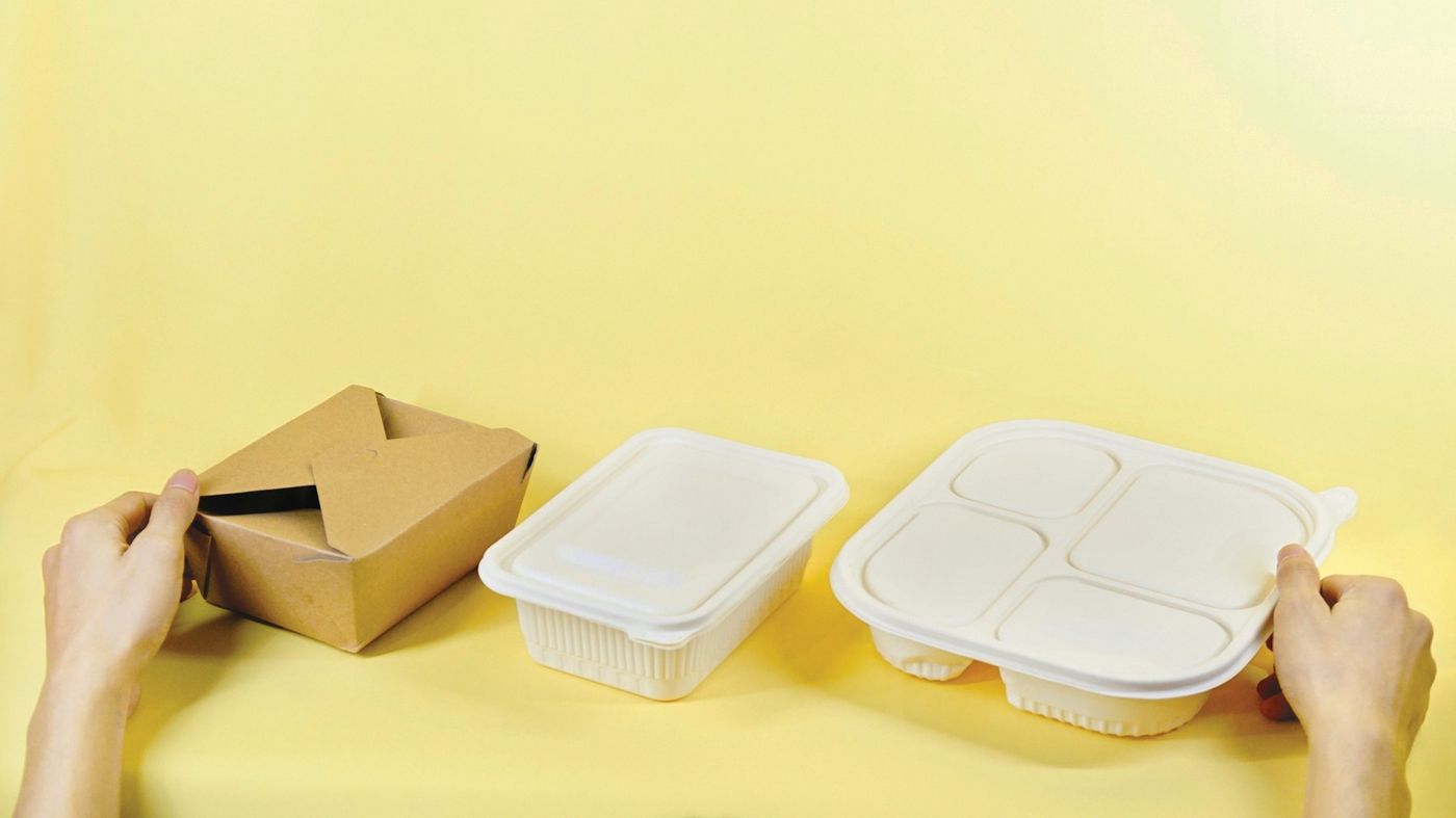 Hands arrange a cardboard takeaway box and two plain food containers on a pale yellow surface
