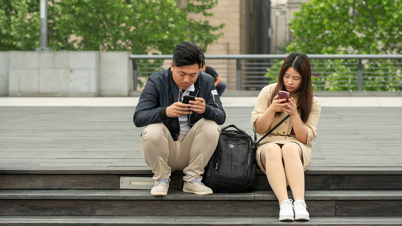Two people sit side by side on outdoor wooden steps, each looking at a phone
