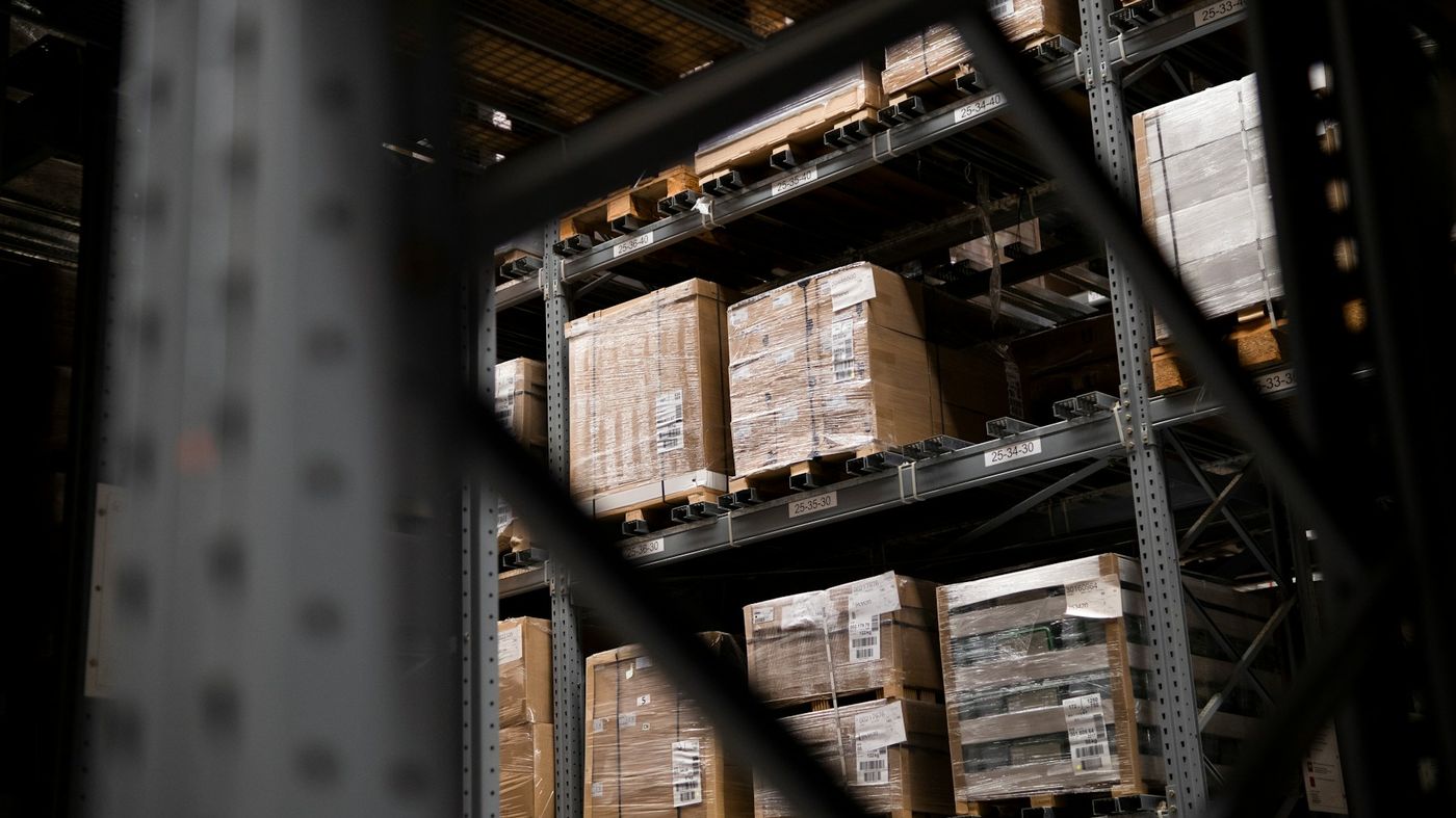 Wrapped pallets of boxes on tall warehouse racking, seen through a steel frame