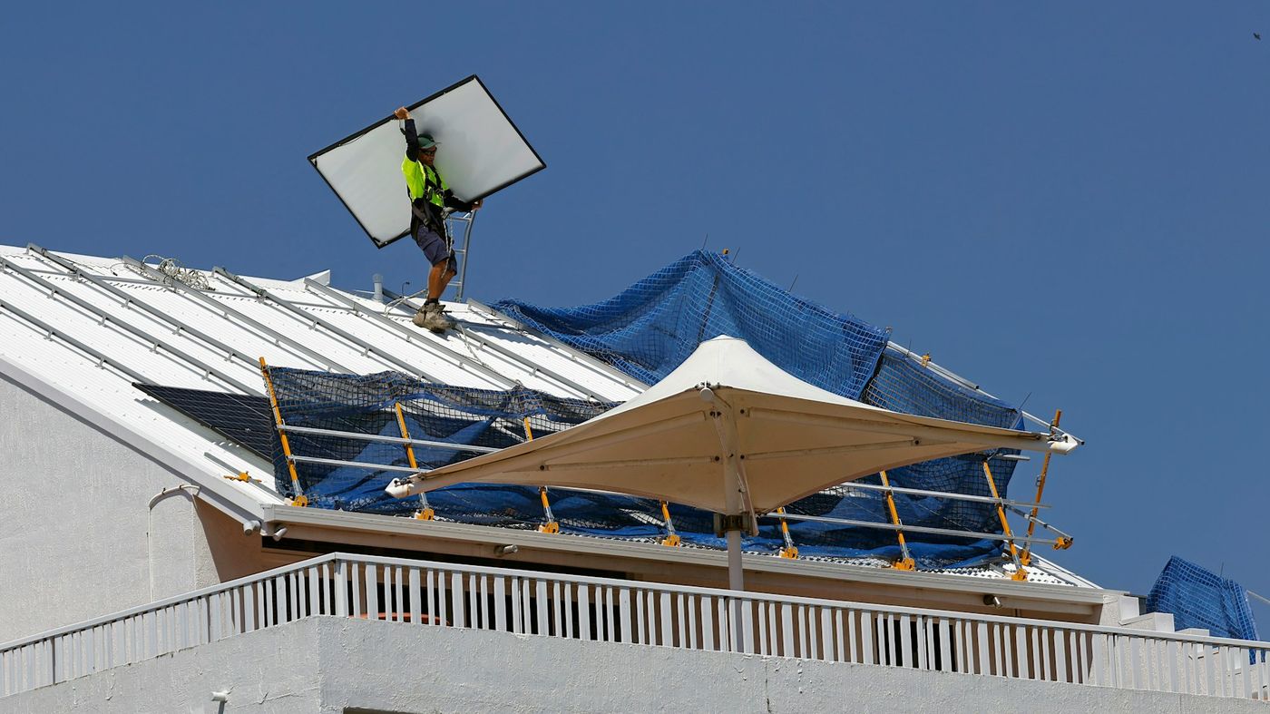 A worker carries a solar panel across a white metal roof under a blue sky