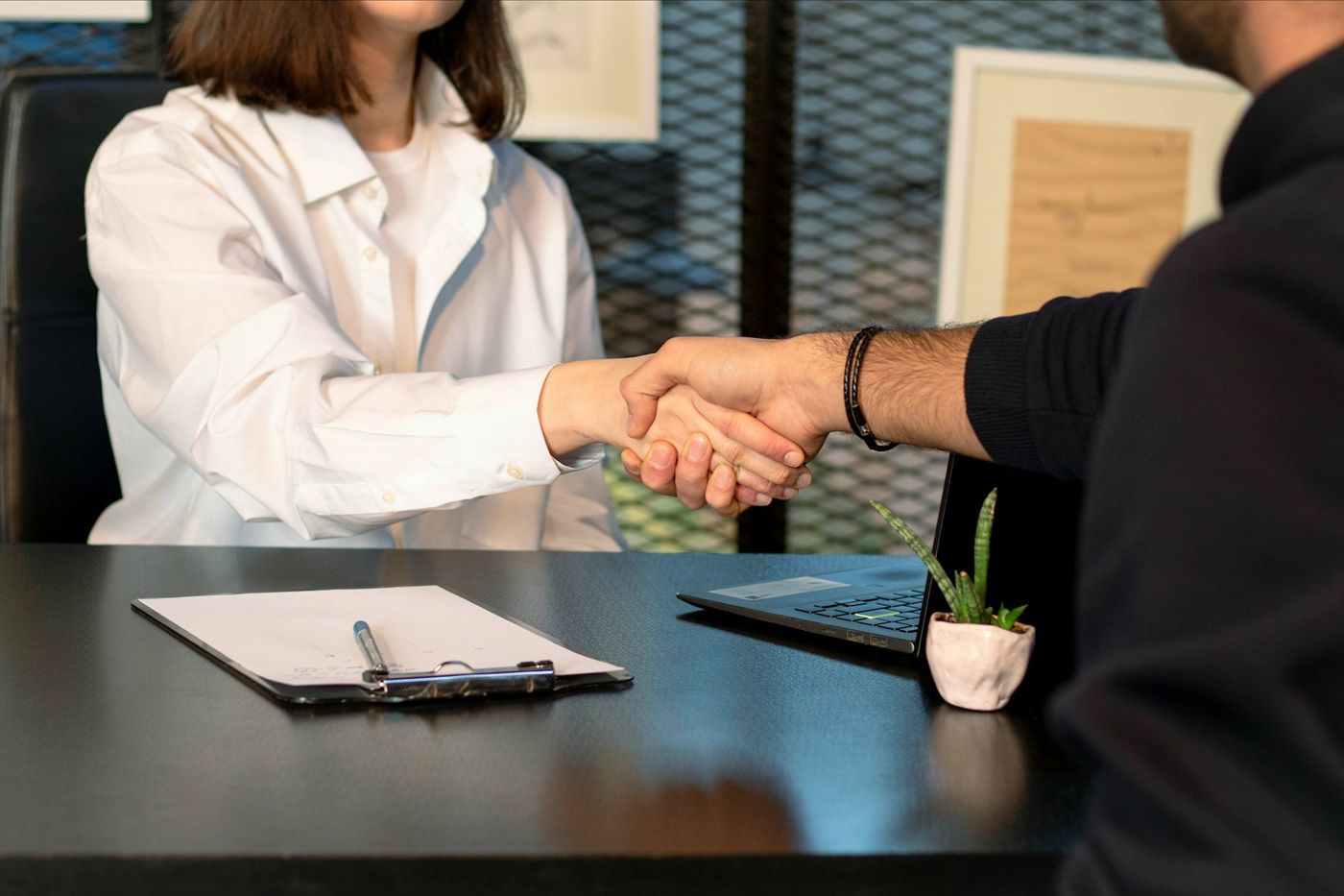 Two people shake hands across a desk with a clipboard and a laptop