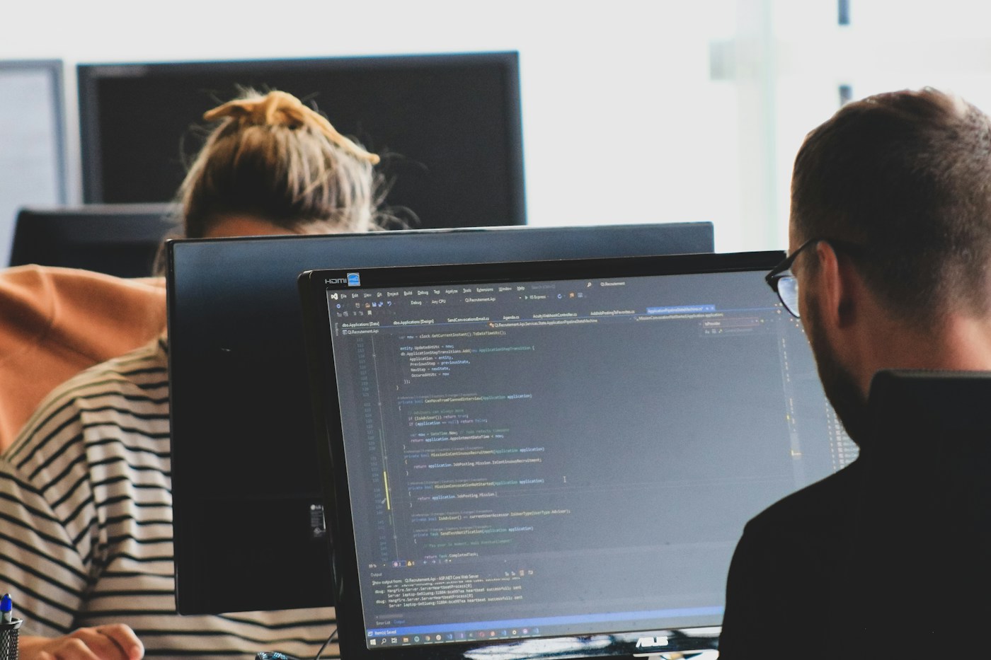 Two people working on code across large monitors in a bright office