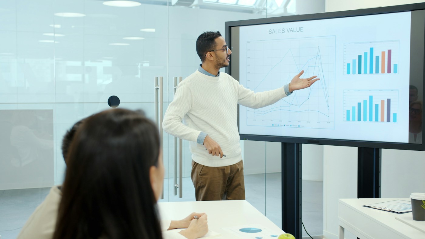 A presenter points to bar charts on a large screen while colleagues watch from the table