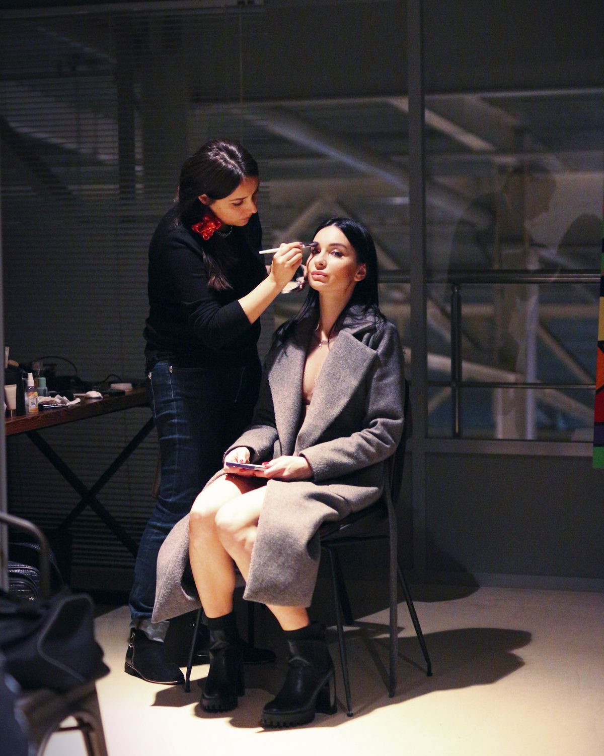 A make-up artist works on a seated model in a dim backstage space