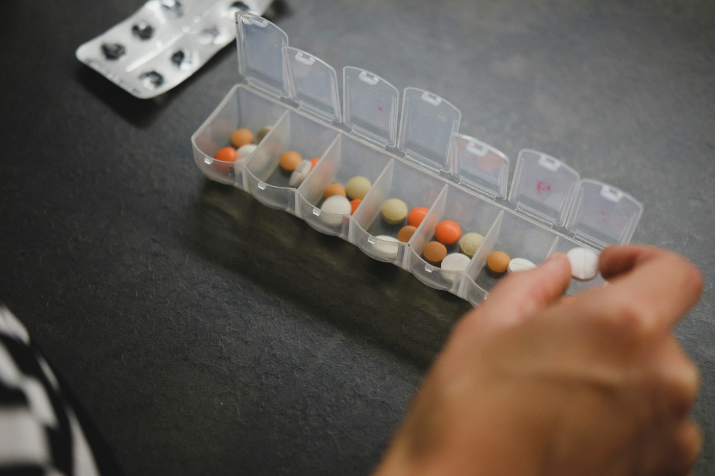A hand taking a tablet from a weekly pill organiser on a dark table