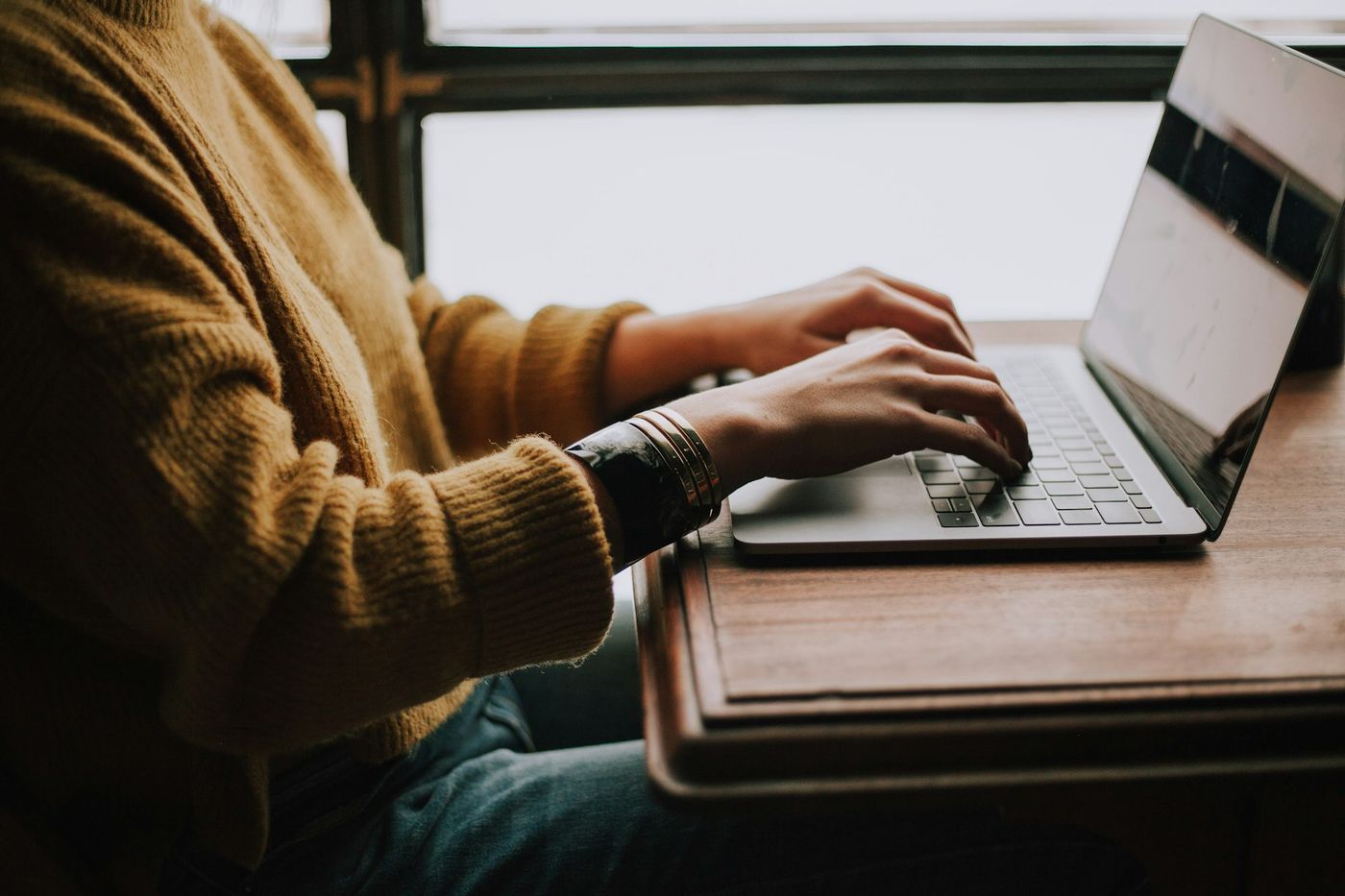 Hands in a mustard sweater type on a laptop resting on a wooden desk