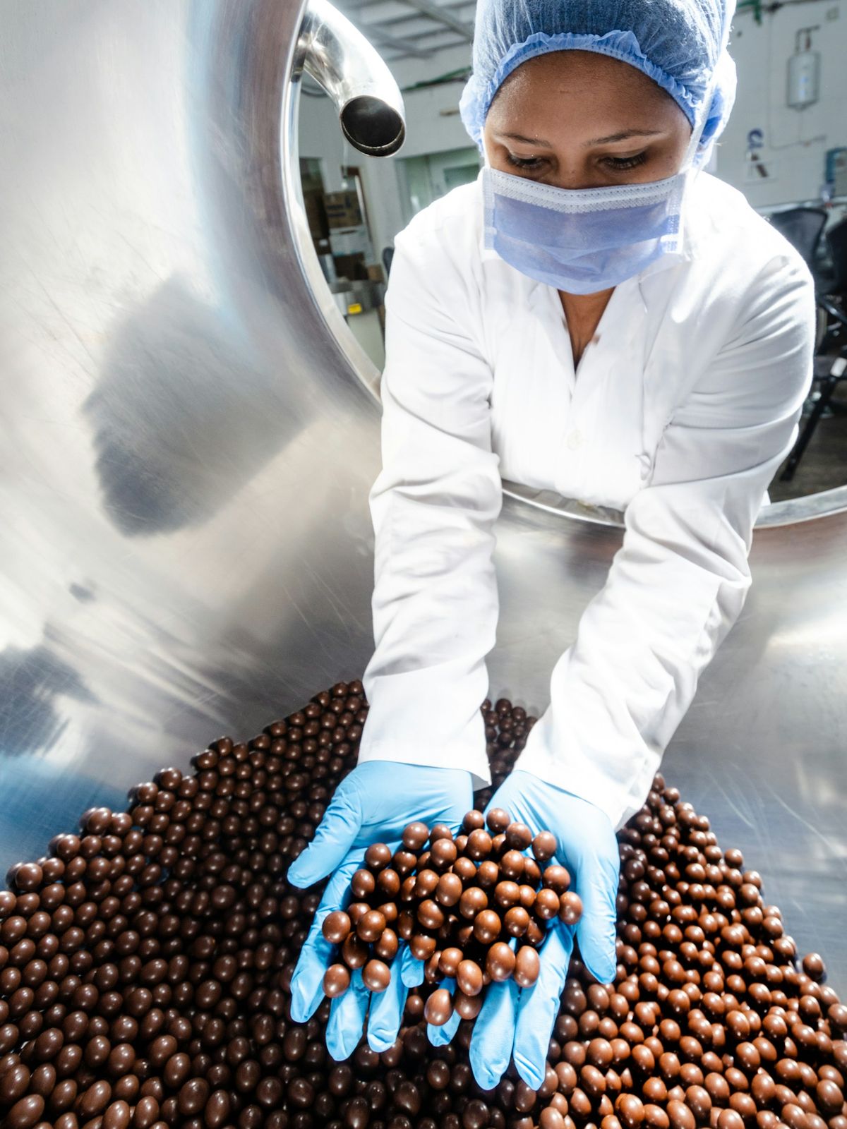 A worker in a hairnet, mask and blue gloves lifts a handful of coated sweets from a polishing pan