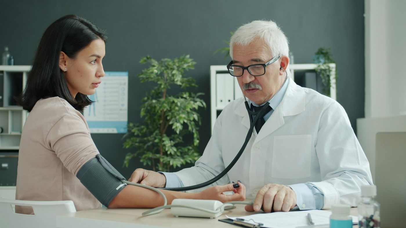 A doctor checks a patient’s blood pressure across a desk in a consulting room
