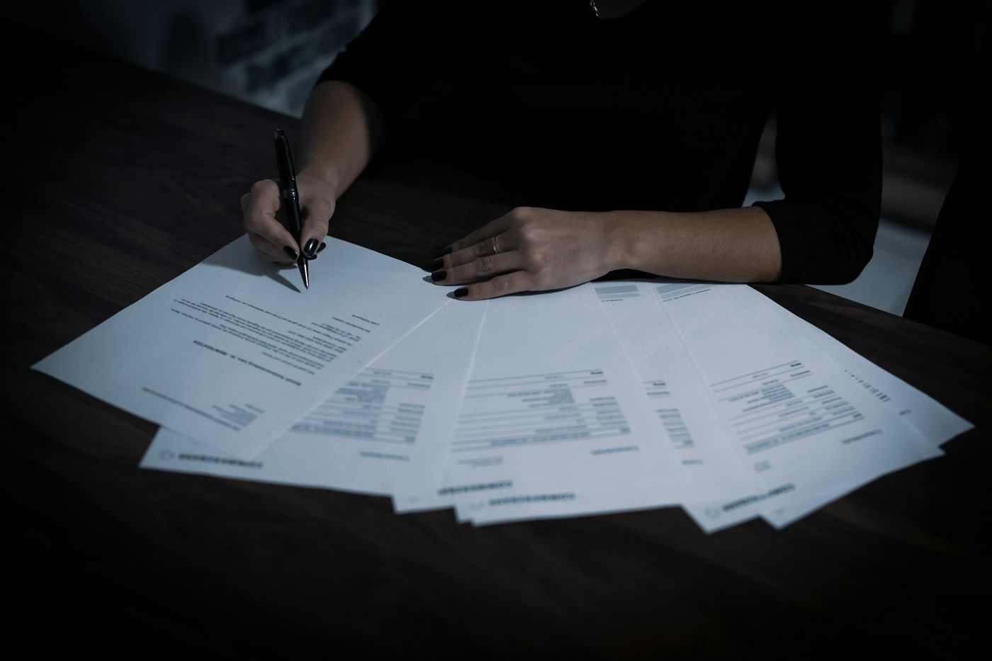 A person signs one of several printed forms spread across a dark table