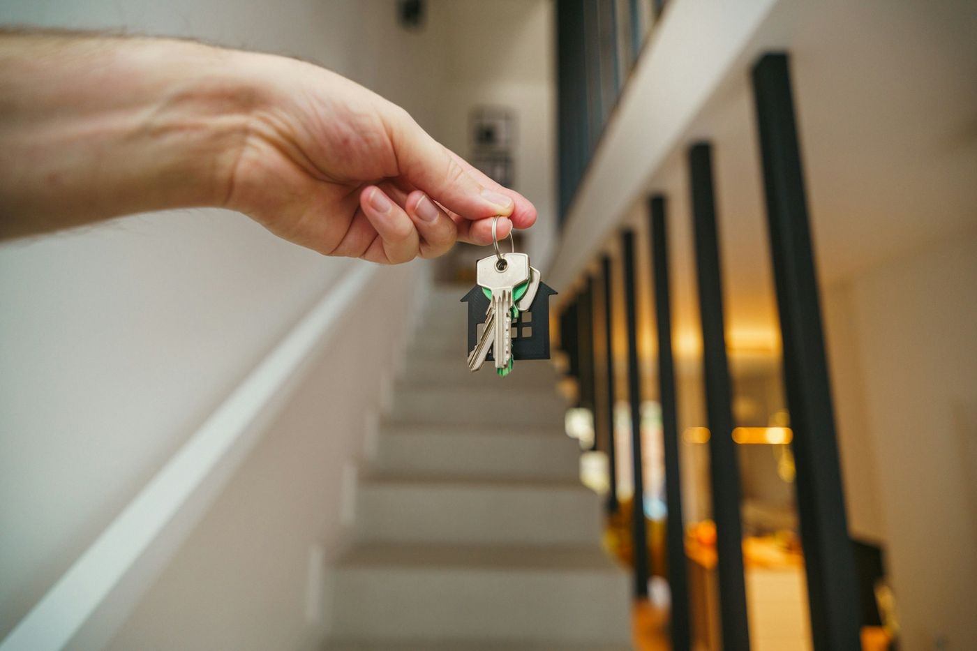 A hand holds a set of keys with a house-shaped key ring beside a staircase