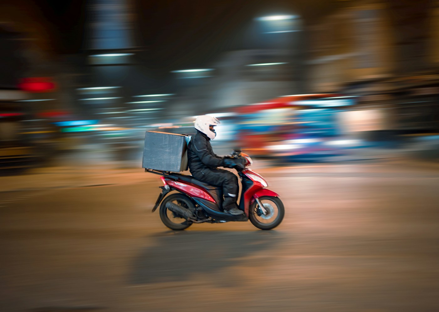 A delivery rider on a red scooter speeds through a city street at night, blurred with motion