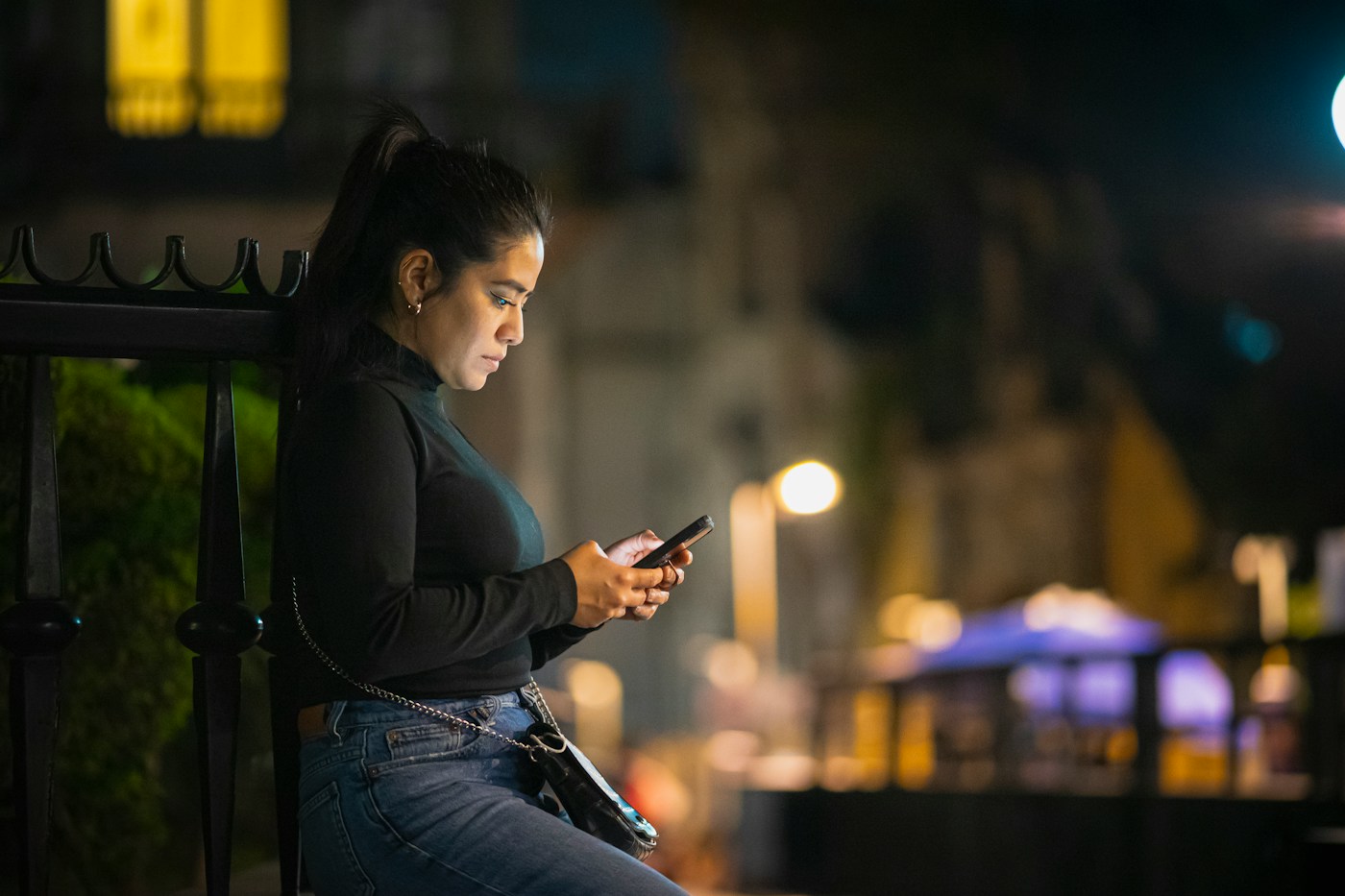 A woman sits on a bench at night looking at her phone, city lights blurred behind her
