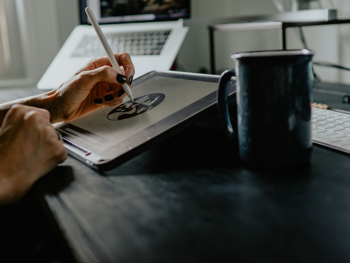 A designer draws on a tablet with a stylus at a desk beside a laptop and a mug