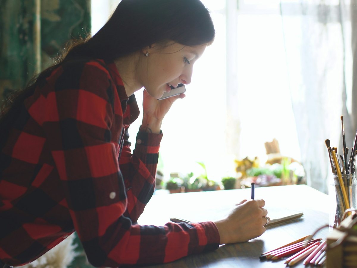 A woman on a phone call writes notes in a notebook at a desk by a bright window