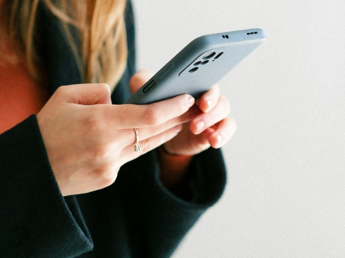 A person in a dark coat holds a phone in both hands against a pale wall