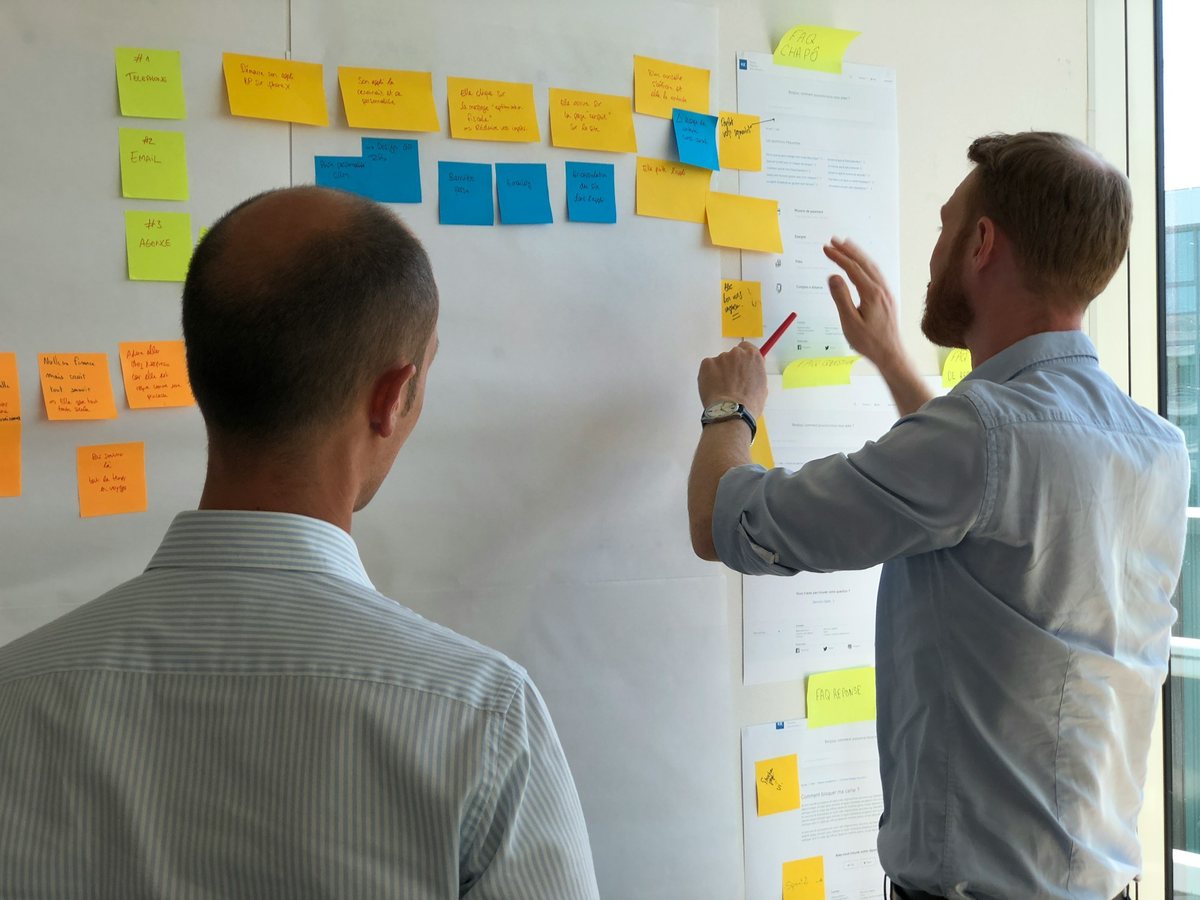 Two colleagues talk through a wall of sticky notes and printed pages